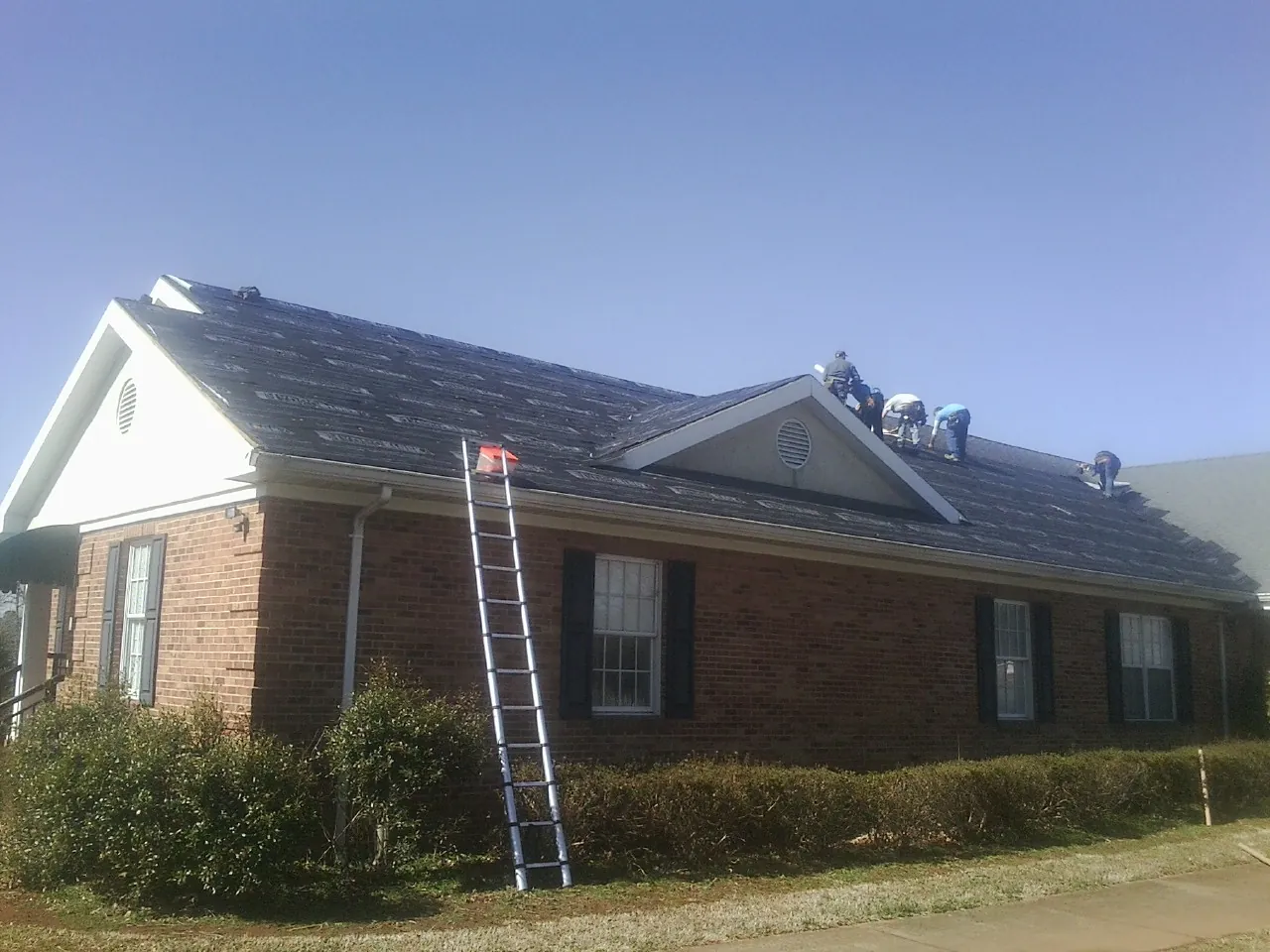 Crew tearing shingles off a brick ranch roof with a ladder set — Metal Roof Repair work by our crew in Burnett