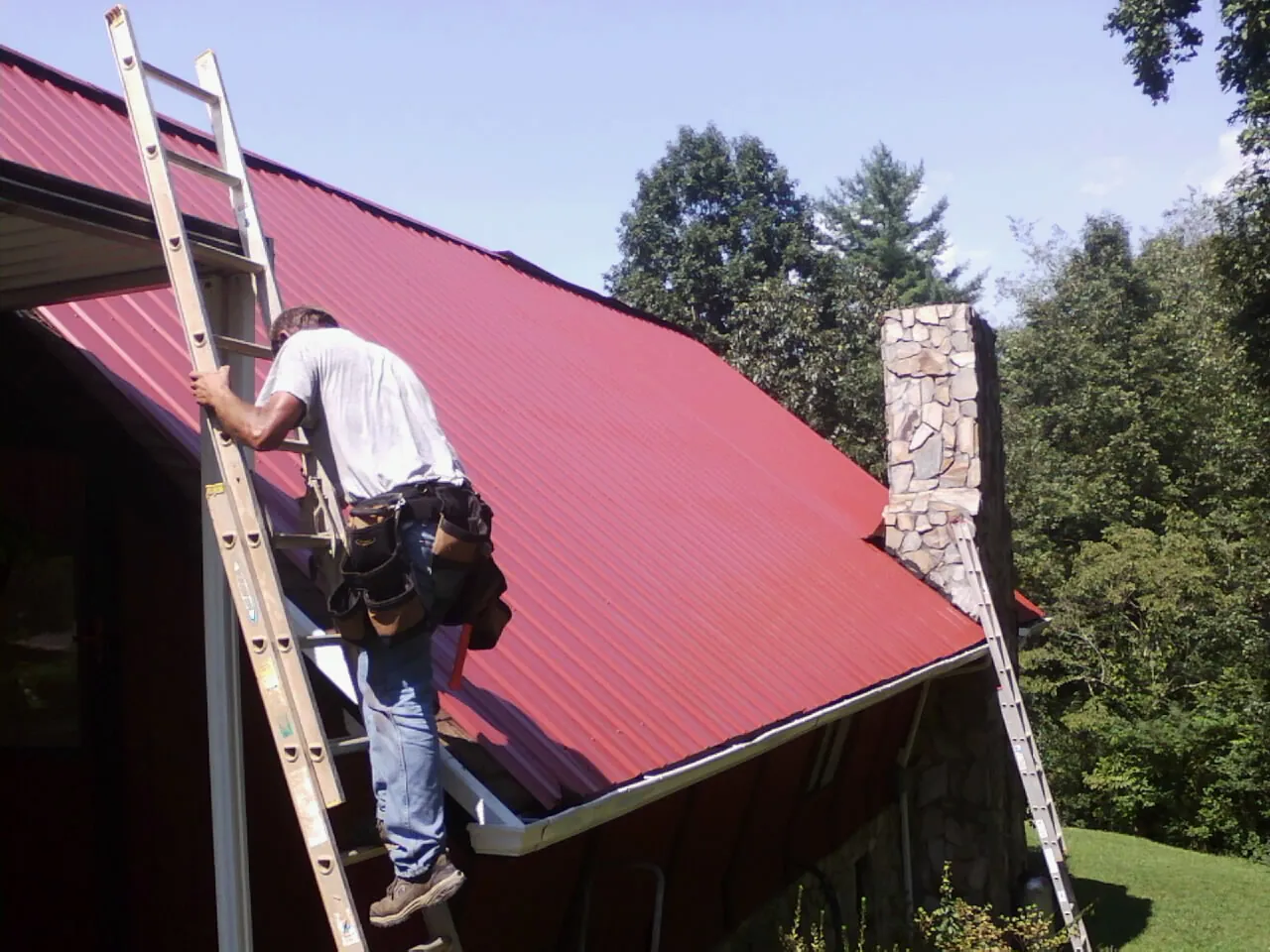 Roofer on a ladder beside a red metal roof and stone chimney — Soffit & Fascia Repair by Roofing Brothers in Burnett